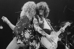 Two rock musicians perform energetically on stage; one sings into a microphone while the other plays an electric guitar. Both have long, curly hair and wear patterned shirts, with stage lighting and a drum set visible in the background.