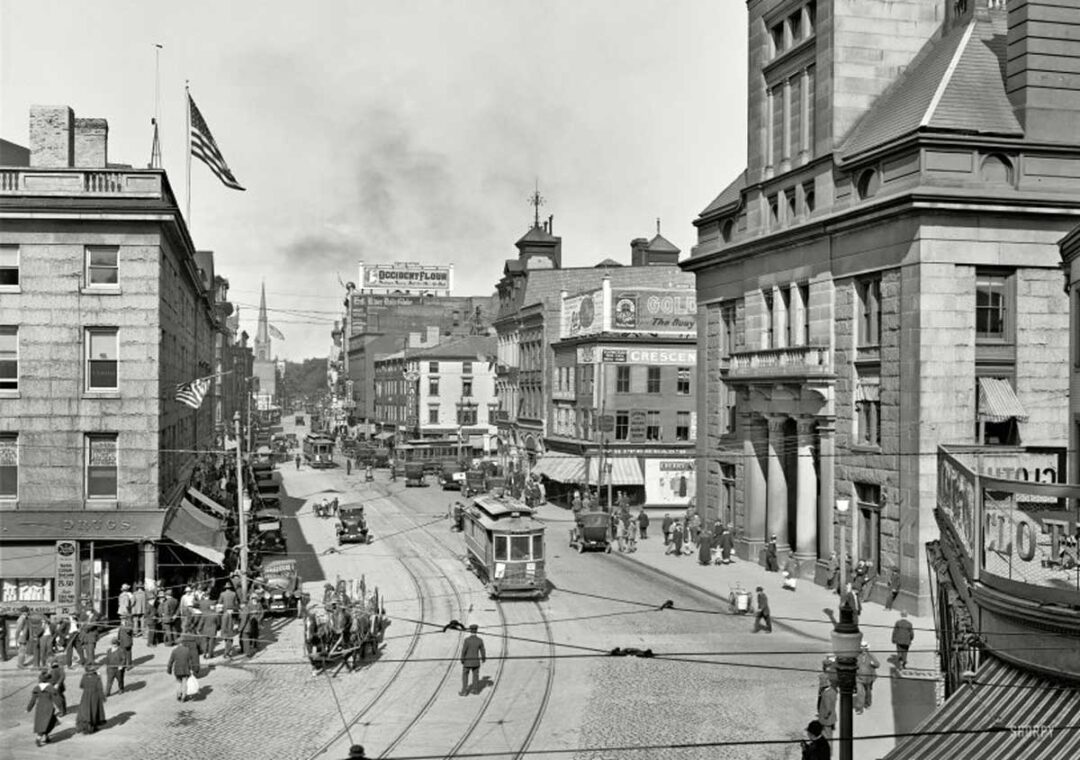 Black and white photo of a busy early 20th-century city street with streetcars, pedestrians, cars, and American flags, surrounded by tall buildings with various shop signs and advertisements.
