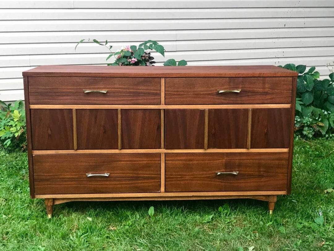 A wooden dresser with six drawers, each featuring a metal handle, stands on a grassy lawn in front of a light-colored siding wall and green plants.