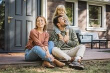 A smiling family of three sits on the front porch of a house. The woman sits cross-legged, the man sits beside her holding a young girl who stands behind him, all appearing happy and relaxed outside their home.