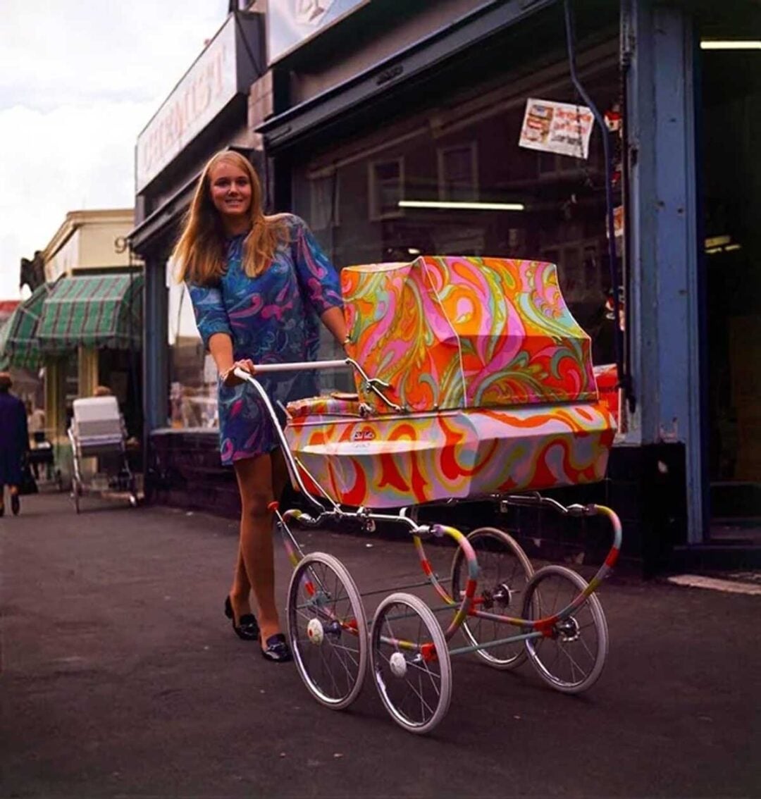 A woman in a colorful dress smiles while pushing a vintage pram with a vibrant, psychedelic pattern on a city sidewalk in front of a store.