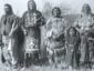 A historical black-and-white photograph of five Native Americans, including three men, one woman, and one young girl, all standing outdoors in traditional clothing and accessories.