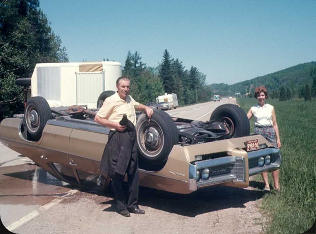 A man and woman stand beside an upside-down car on the side of a rural road. The man holds a jacket and hat, while the woman smiles. Trees and vehicles are visible in the background.
