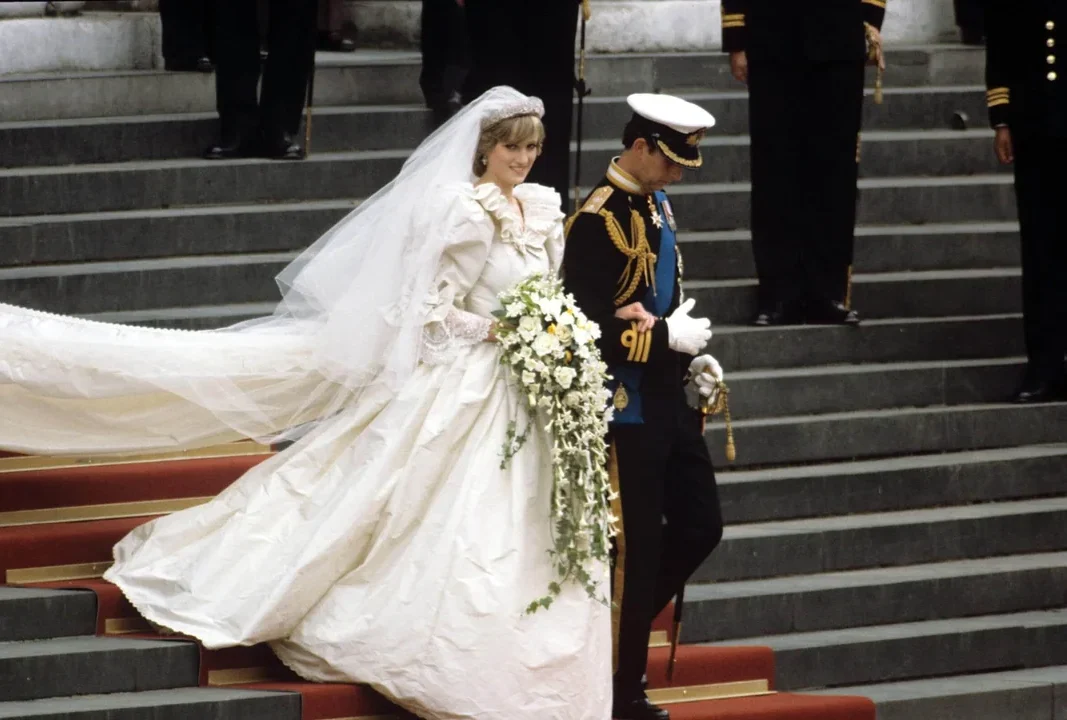 A bride in a white wedding gown and veil walks down red-carpeted steps, holding a cascading bouquet. She is escorted by a man in a formal military uniform. People in dark suits stand in the background.