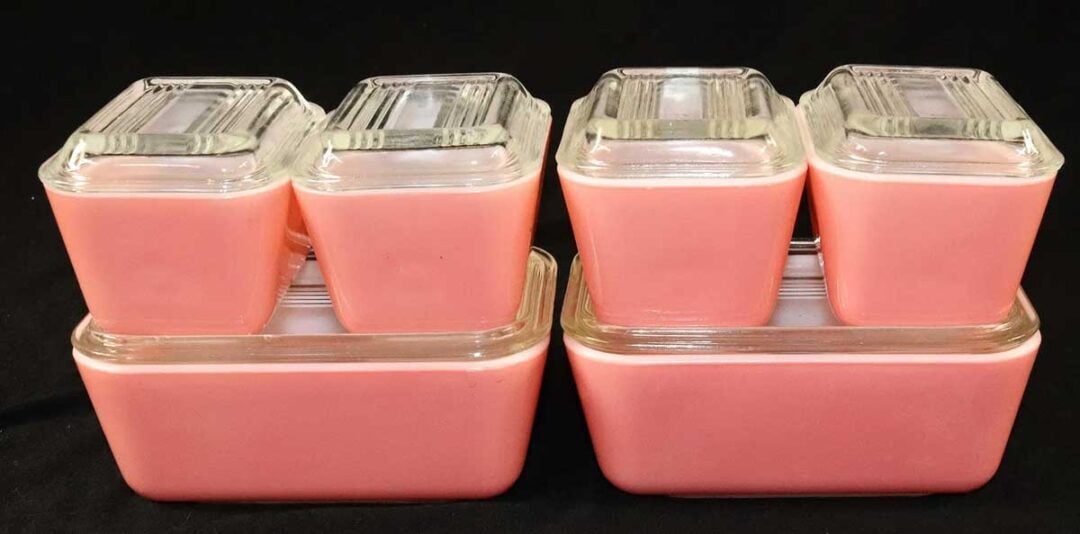 Four vintage pink glass refrigerator dishes with clear glass lids are stacked in two sets against a black background. Each set has two small dishes on top of one larger dish.