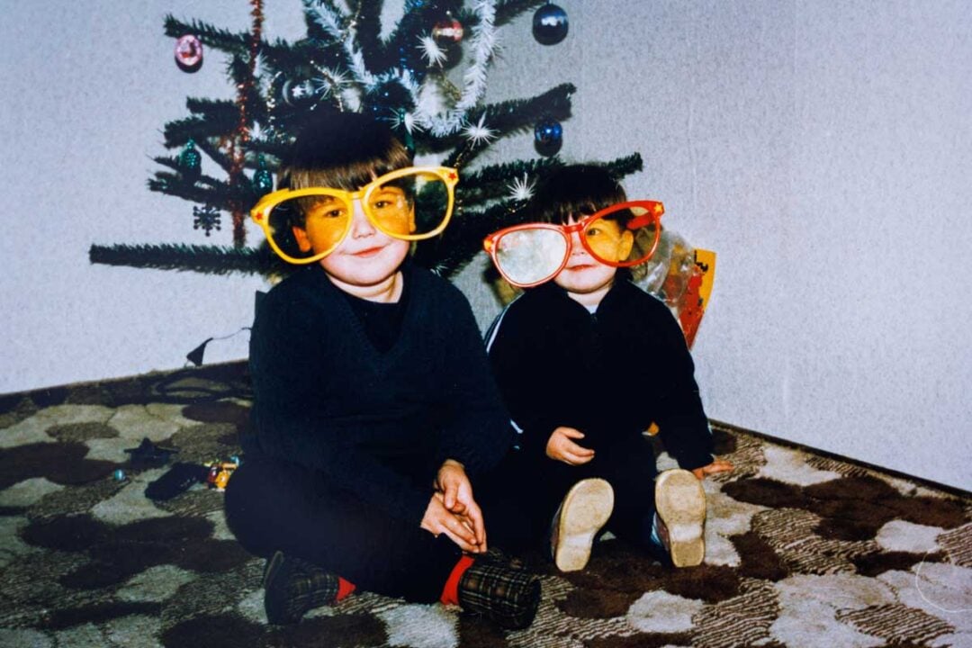 Two young children wearing oversized, colorful novelty glasses sit on a patterned carpet in front of a decorated Christmas tree, smiling and posing for the camera.