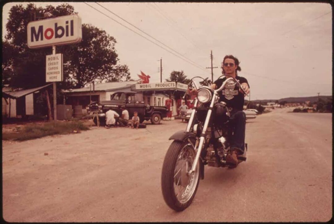 A man rides a motorcycle down a rural road past a vintage Mobil gas station, where people are gathered near old vehicles. The scene appears to be set in the 1970s.