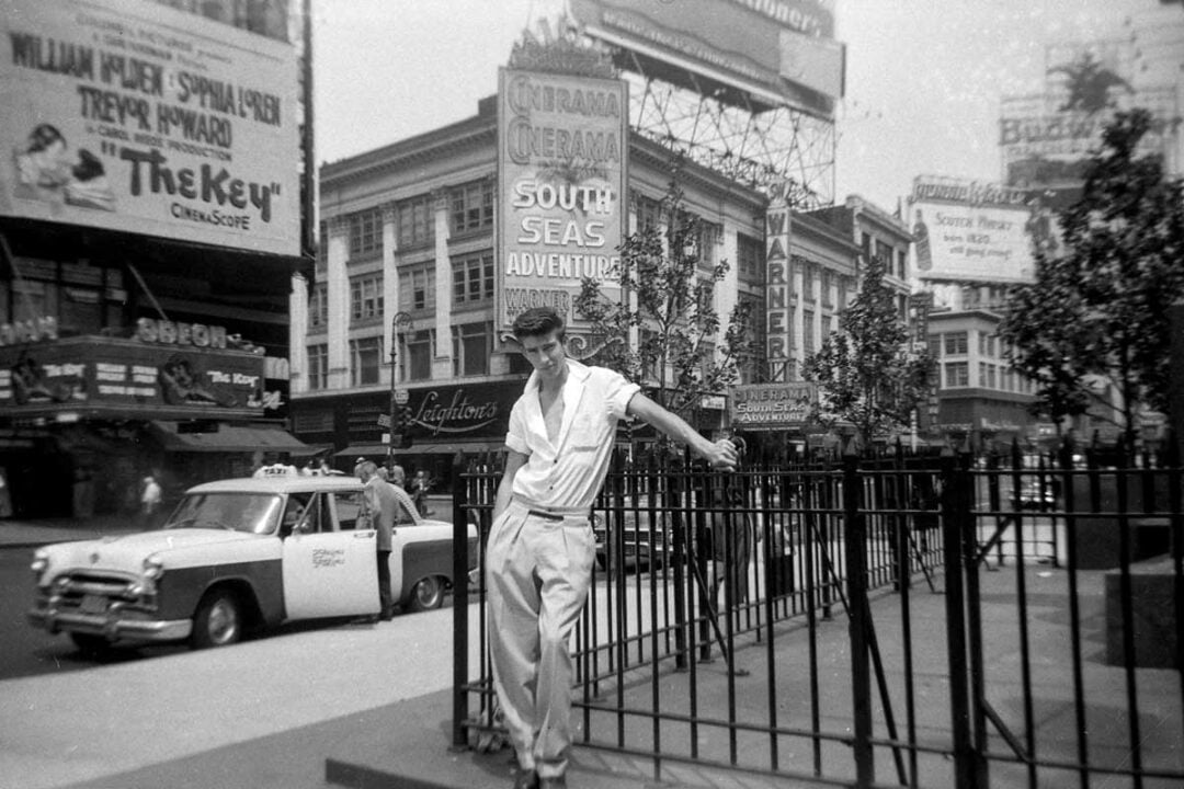 A young man in casual clothes leans on a metal fence in a busy city street with vintage cars, billboards, and old movie theater signs in the background. The scene appears to be from the mid-20th century.