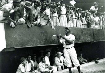A baseball player in uniform stands below a stadium fence, signing autographs for a crowd of eager fans leaning over the railing. Teammates and coaches sit on a bench in the dugout behind him. The image is black and white.