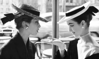 Two women in elegant hats and pearl earrings sit across from each other at a caf&eacute;. One holds a pearl necklace, displaying it on her hand, while the other looks at it intently. The scene is in black and white.
