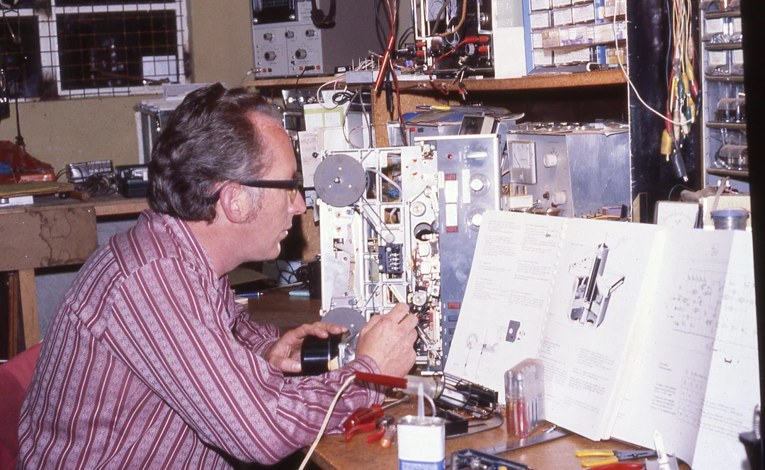 A man in glasses and a striped shirt sits at a cluttered workbench, repairing electronic equipment, with open technical diagrams and tools scattered around him.