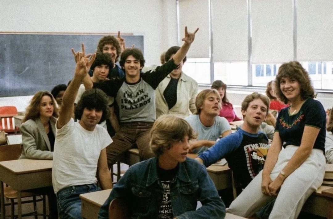 A group of teenagers smiling and posing in a classroom, some making hand gestures. Desks, chairs, and a chalkboard are visible in the background. The mood is cheerful and friendly.