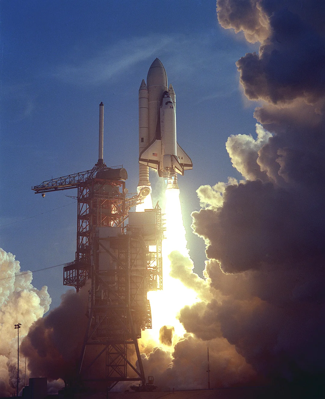 A space shuttle launches from a pad, with bright flames and smoke billowing underneath as it ascends into a clear sky, surrounded by large clouds of exhaust.