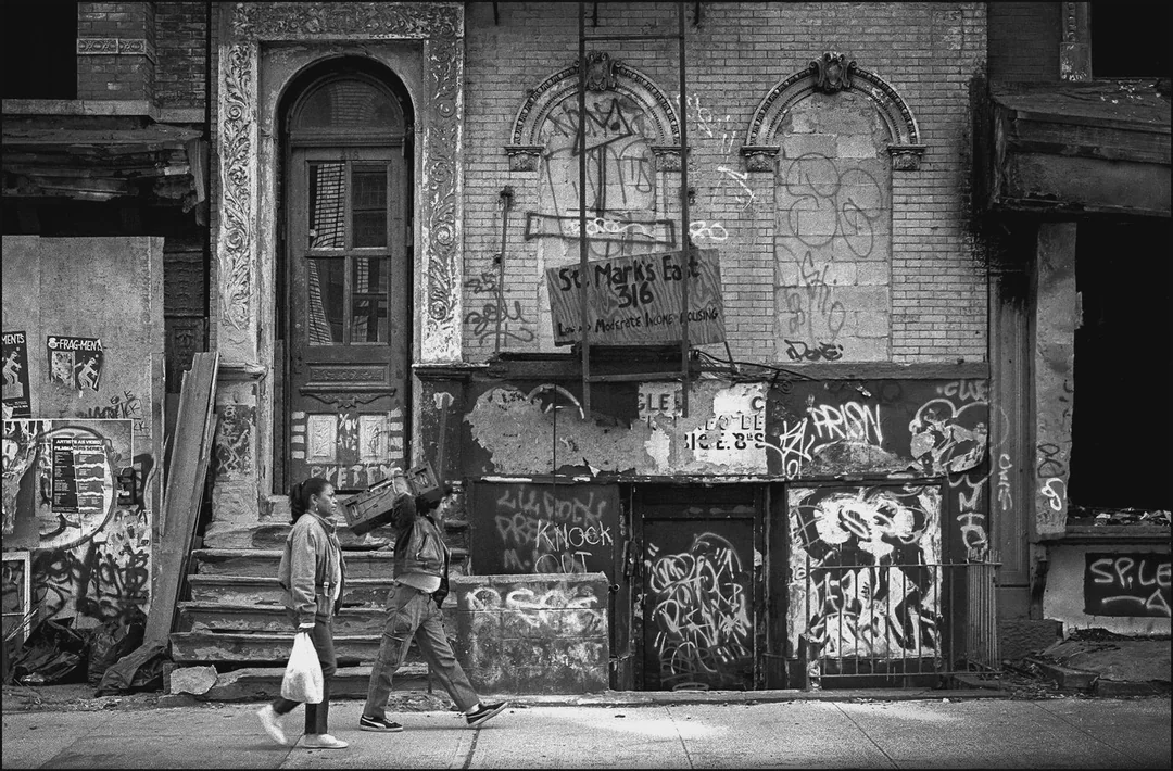 Two people walk past a rundown, graffiti-covered building with boarded-up windows and doors, surrounded by urban decay. One person carries a bag, and the scene has a gritty, neglected city atmosphere.