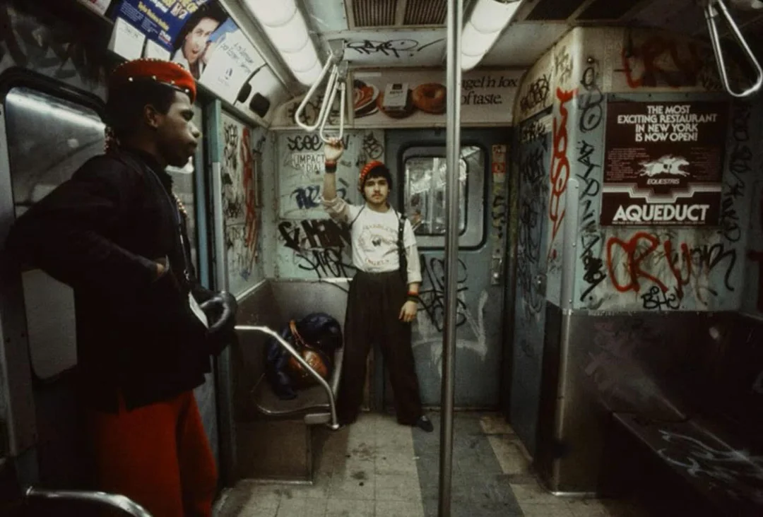 Two men stand in a graffiti-covered subway car; one leans against the door wearing a red beret and gloves, while the other stands by the opposite door, both surrounded by worn seats and dim lighting.