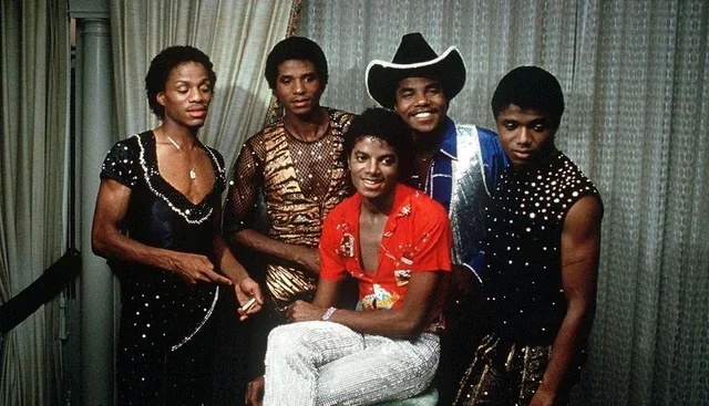 Five men dressed in colorful, sequined stage outfits pose together indoors, smiling and standing in front of draped curtains. One man in a red shirt sits while the others stand around him.