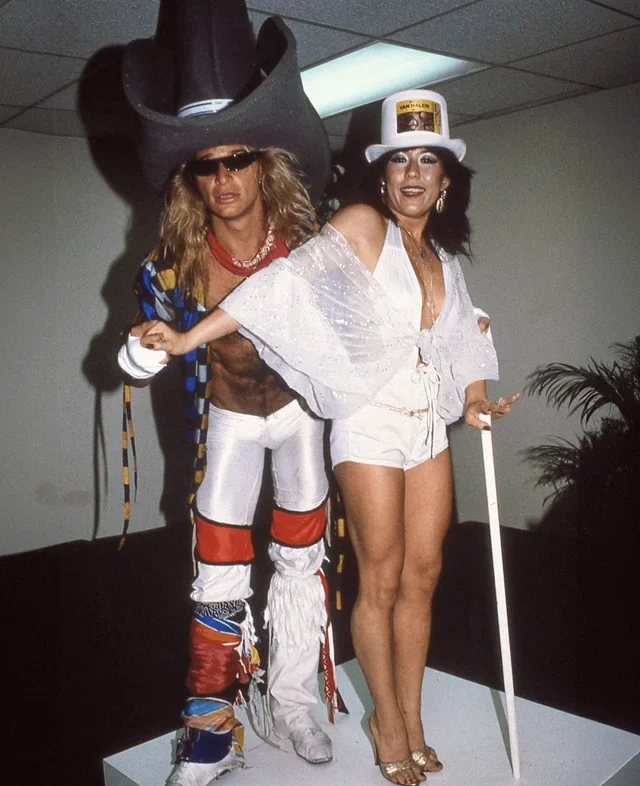 A man and woman in eccentric costumes pose on a white platform. The man wears a giant black cowboy hat, sunglasses, and fringed boots, while the woman wears a white top hat, sheer cape, white outfit, and holds a white cane.