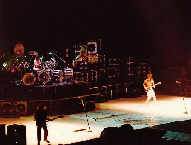 A rock band performs on a large stage with stacks of speakers and bright lighting. The drummer plays on a raised platform while two guitarists stand spread apart on the stage. The scene is energetic and dramatic.