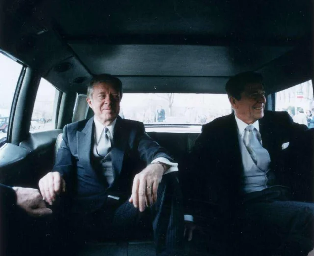 Two men in suits sit in the back seat of a car, facing forward and smiling. The interior is dimly lit, and daylight is visible through the windows behind them.
