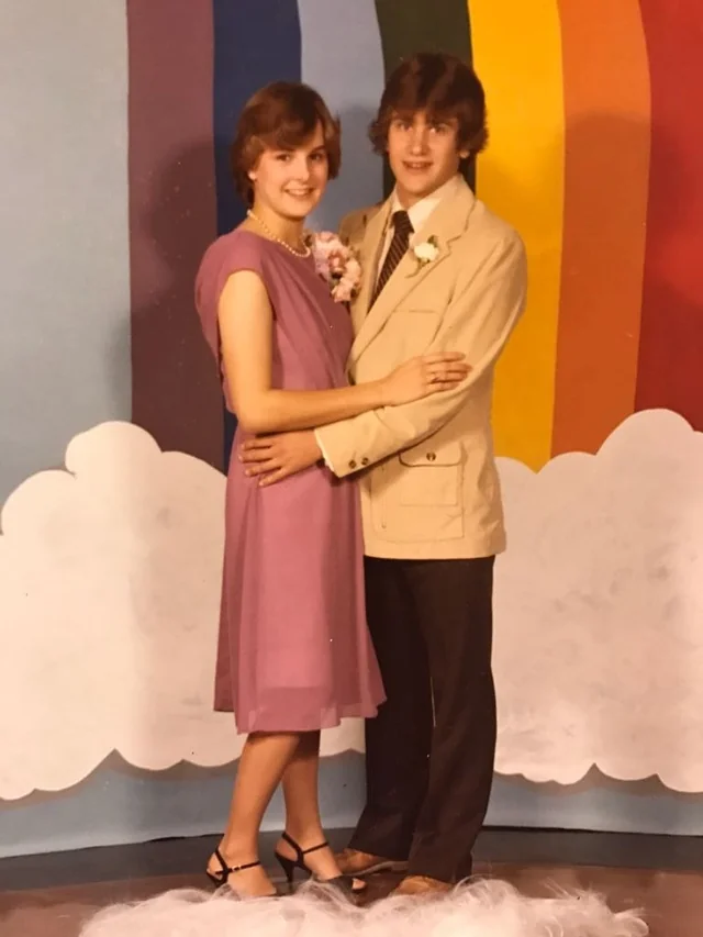 A young couple poses together in front of a backdrop with clouds and rainbow-colored stripes. The girl wears a purple dress and heels, while the boy wears a beige suit and brown shoes. Both wear corsages and smile at the camera.