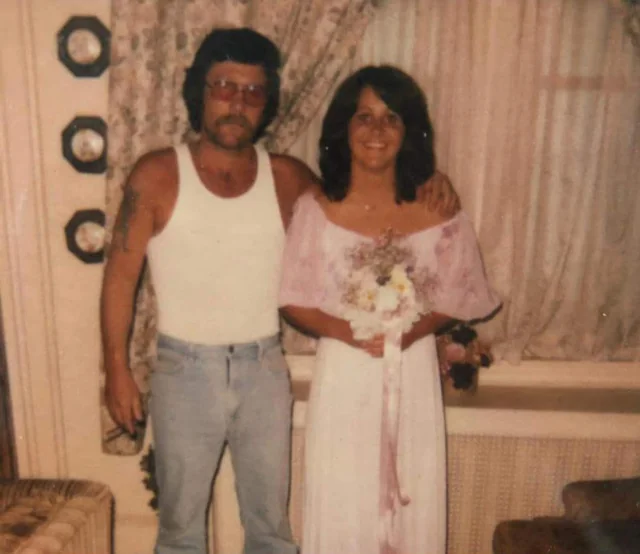 A man in a white tank top and jeans stands beside a woman in a pink dress holding a bouquet, both smiling in a living room with curtains and decorative plates on the wall.
