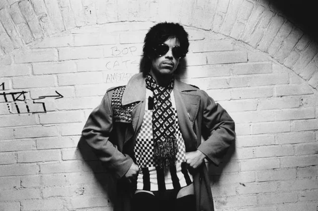 A person wearing sunglasses, a patterned scarf, and a long coat stands against a white brick wall with hands on hips. Graffiti and an arrow are visible on the wall behind them.
