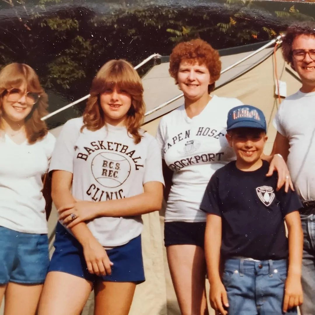 Five people, three women, a young boy, and a man, stand outside in front of a tent, smiling. They are casually dressed in shorts and t-shirts, and trees are visible in the background.
