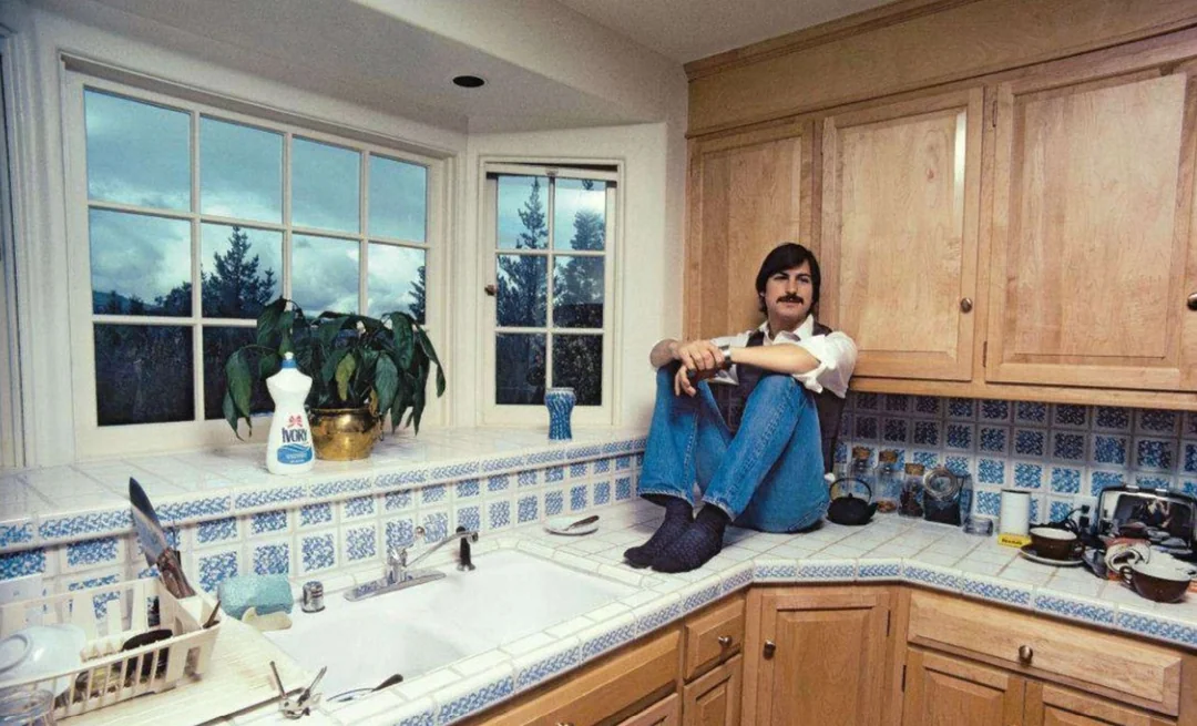 A man with dark hair and a mustache sits barefoot on a kitchen counter, knees drawn to his chest, beside a sink and window with a view of trees and sky; various dishes and plants are visible on the countertop.