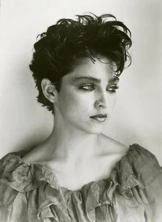 Black and white portrait of a young woman with short, tousled dark hair, wearing a textured, off-the-shoulder top, looking pensively to the side against a plain background.