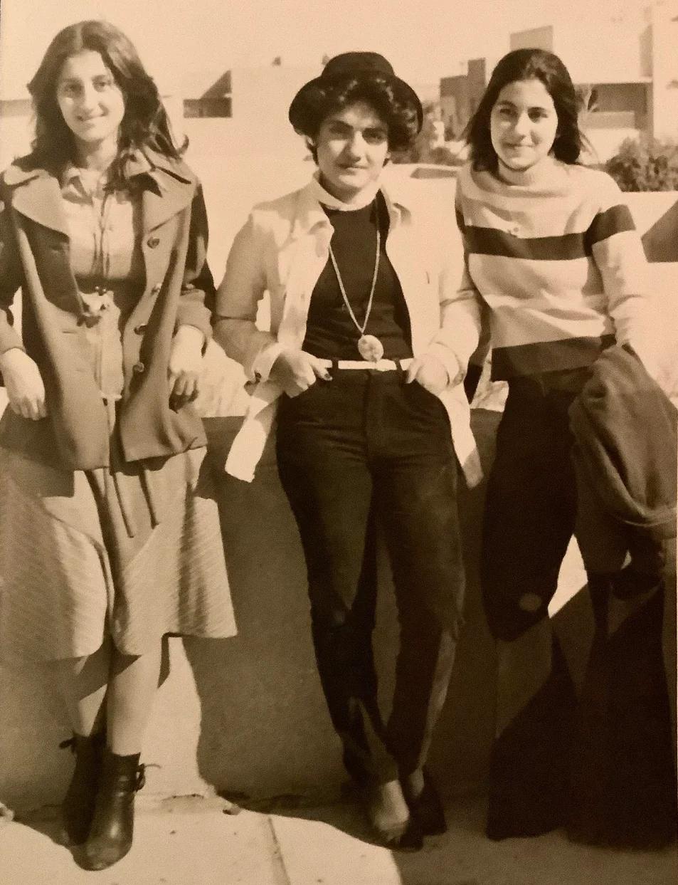 Three young women in vintage clothing pose together outdoors against a low wall. Two are standing on either side of the third woman, who is leaning back and wearing a hat and long necklace. The photo is black and white.