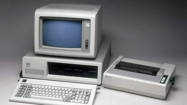 An old IBM personal computer with a CRT monitor, floppy disk drives, a keyboard, and a dot matrix printer, all in beige, displayed on a gray surface.