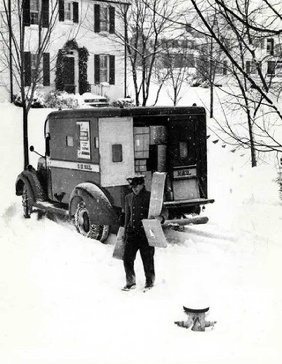 A postal worker carries packages through deep snow toward a house, with a vintage U.S. mail truck parked nearby on a snowy street. Snow covers the ground and trees, and houses are visible in the background.