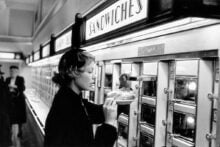A woman in a coat selects food from a self-serve automat, opening a small glass door labeled "Sandwiches" to retrieve a plate, with more food compartments visible along the wall. The scene is black and white.