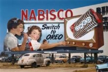 A vintage billboard advertises Nabisco Oreo cookies, showing two girls eating Oreos and a hand reaching for an Oreo from a TV screen. The sign reads &ldquo;Switch to OREO.&rdquo; Classic cars are parked below.