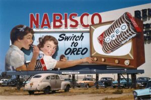 A vintage billboard advertises Nabisco Oreo cookies, showing two girls eating Oreos and a hand reaching for an Oreo from a TV screen. The sign reads “Switch to OREO.” Classic cars are parked below.