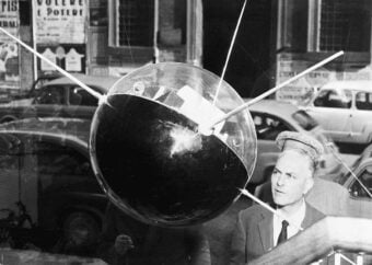 A man in a suit stands near a large model of the Sputnik satellite, displayed in a window with cars and posters visible on the street outside.