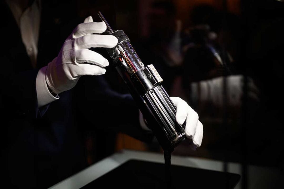 A person wearing white gloves carefully holds a metallic, futuristic object resembling a lightsaber, displayed under focused lighting against a dark background.