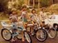 Four children stand with their colorful vintage bicycles on a suburban street, smiling and barefoot, with houses, trees, and a white car in the background. It appears to be a warm, sunny day.
