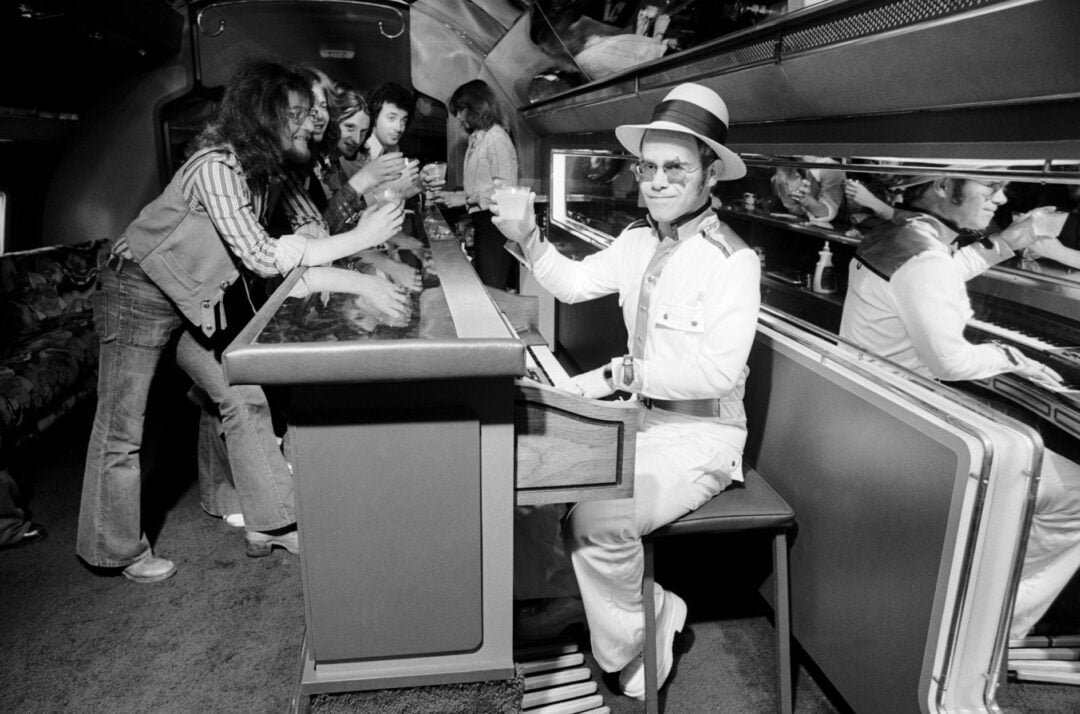 A man in a white suit and hat sits at a bar on a train or plane, raising a glass and smiling, while several casually dressed people stand and interact at the bar in the background.