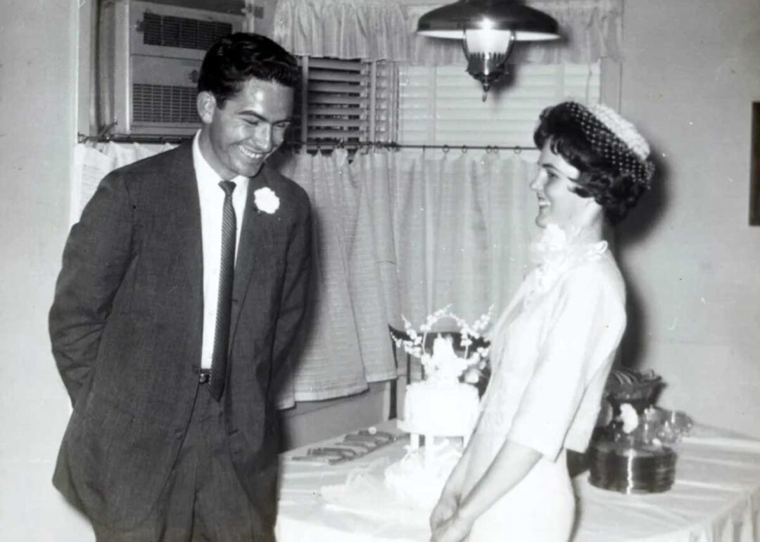 A man in a suit and a woman in a dress and veil smile at each other in front of a decorated cake, possibly at a wedding celebration, in a room with curtains and an air conditioner in the background.