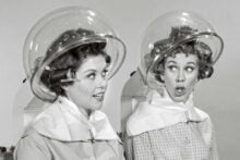 Two women with curlers in their hair sit under vintage hair dryers at a salon, wearing capes and chatting; one looks surprised while the other smiles. The image is in black and white.