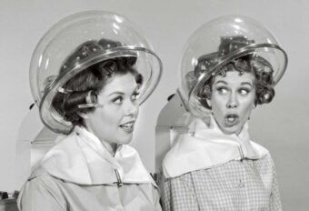Two women with curlers in their hair sit under vintage hair dryers at a salon, wearing capes and chatting; one looks surprised while the other smiles. The image is in black and white.