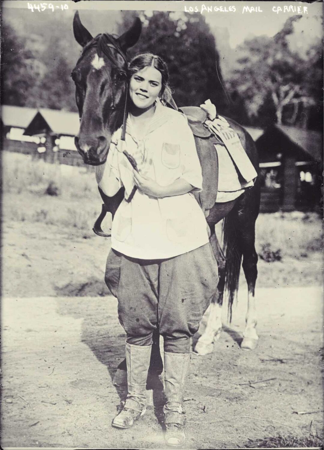 A smiling woman in riding clothes stands beside a saddled horse, holding its head gently. Wooden buildings and trees are visible in the background. The top of the image reads "LOS ANGELES MAIL CARRIER.