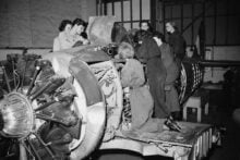 A group of women in coveralls work together on a large aircraft engine inside an industrial workshop, examining parts and making adjustments.