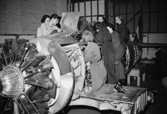 A group of women in coveralls work together on a large aircraft engine inside an industrial workshop, examining parts and making adjustments.