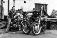 Four women in leather jackets pose confidently with two motorcycles in front of a vintage car and a brick wall, evoking a rebellious, 1970s biker gang vibe.