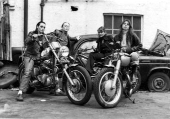 Four women in leather jackets pose confidently with two motorcycles in front of a vintage car and a brick wall, evoking a rebellious, 1970s biker gang vibe.