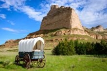 A covered wagon sits on green grass in front of a large rocky butte under a blue sky with scattered clouds. Shrubs and small trees grow near the base of the butte.