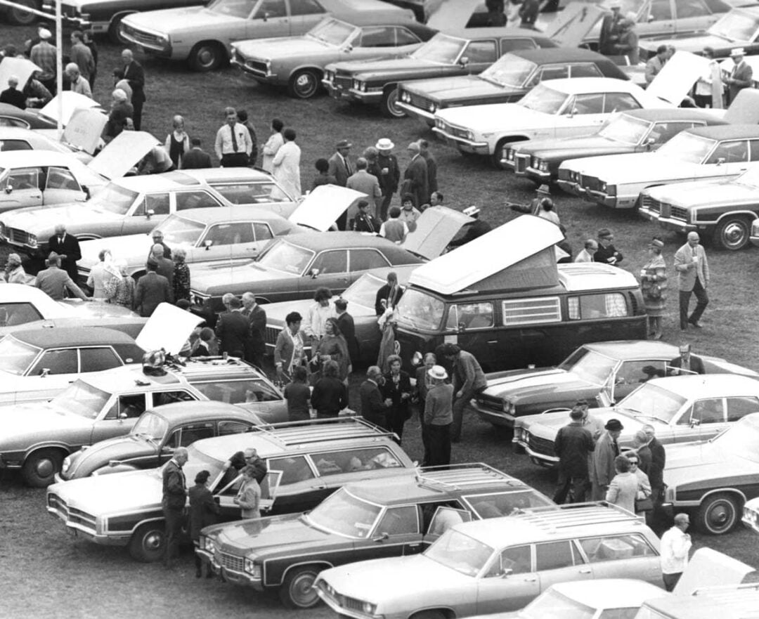 Black and white photo of a crowded car lot with people gathered around cars, many with open hoods. Most vehicles are station wagons and vintage sedans; one van with a raised roof is in the center. The event appears busy and social.