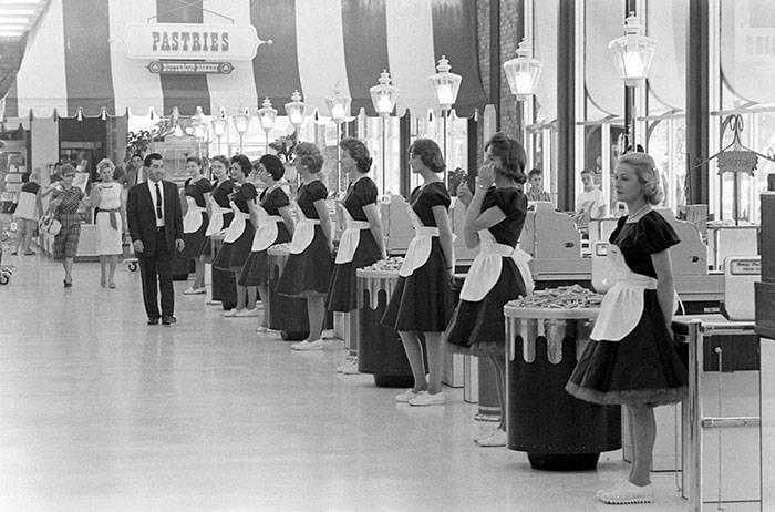 A row of female cashiers in matching uniforms and aprons stand beside checkout counters in a grocery store, while a man in a suit walks by. Customers shop in the background near a sign that reads "Pastries.
